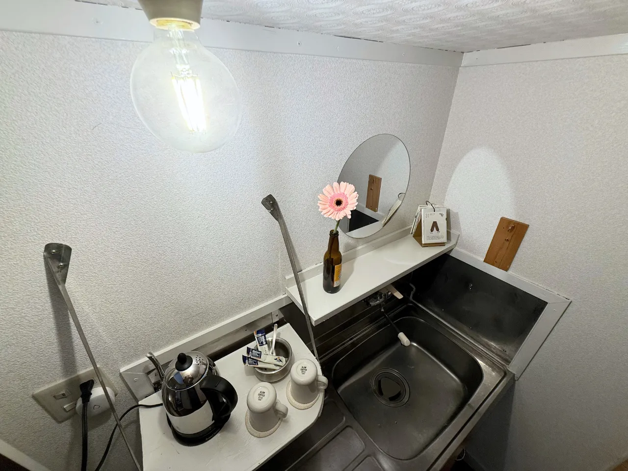 共有キッチン。電気ケトルとコーヒーカップ、シンクの上に丸い鏡と一輪の花